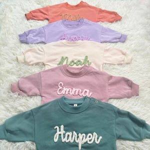 May include: Four pastel-colored baby sweatshirts with embroidered names: Mila, Avery, Noah, Emma, and Harper. The sweatshirts are in shades of coral, lavender, cream, pink, and teal. Each has a crew neck and snap closure on the shoulder.