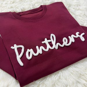 May include: A maroon sweatshirt with the word "Panthers" in white, textured lettering. The crew neck sweatshirt is folded on a white, fluffy surface. The text is in a cursive style.