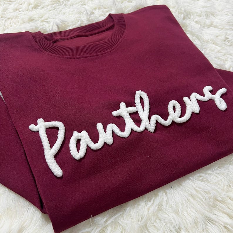 May include: A maroon sweatshirt with the word "Panthers" in white, textured lettering. The crew neck sweatshirt is folded on a white, fluffy surface. The text is in a cursive style.