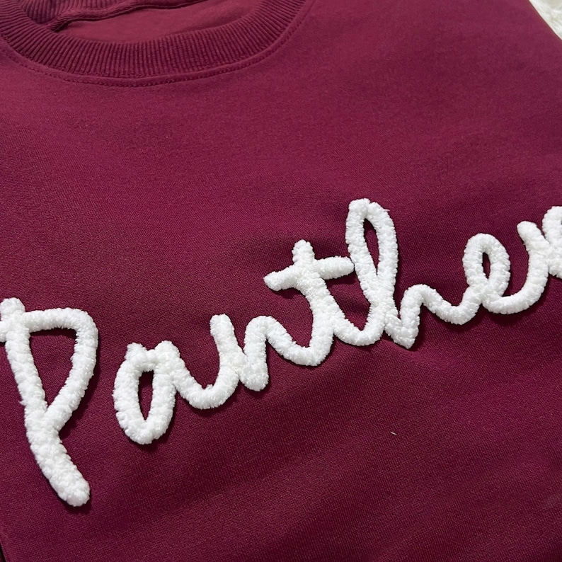 May include: A maroon sweatshirt with the word "Panther" embroidered in white, fluffy, cursive lettering. The sweatshirt has a crew neck and a simple, clean design, perfect for casual wear.