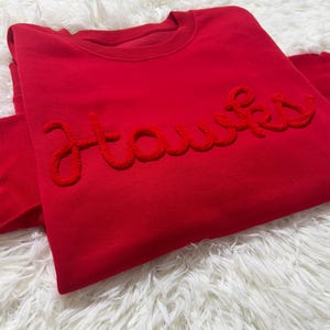 May include: A red sweatshirt featuring the word "Hawks" in a textured, red embroidery. The garment is folded on a white, fluffy surface.