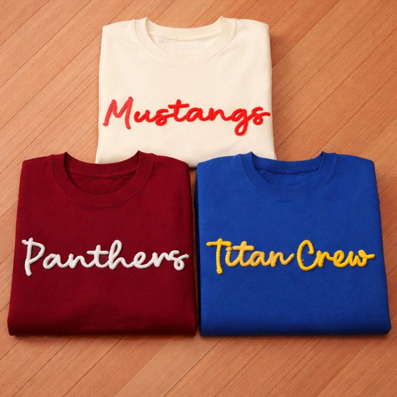 May include: Three folded sweatshirts in cream, burgundy, and blue. Each shirt features a team name in a contrasting color: "Mustangs" in red, "Panthers" in white, and "Titan Crew" in yellow. The shirts are displayed on a wooden surface.