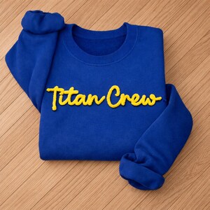 May include: A royal blue sweatshirt with the text "Titan Crew" in yellow script. The sweatshirt is folded, with the sleeves cuffed. The fabric appears soft and comfortable, suitable for casual wear.