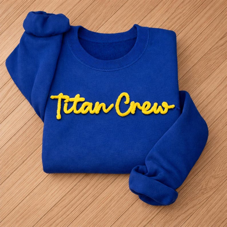 May include: A royal blue sweatshirt with the text "Titan Crew" in yellow script. The sweatshirt is folded, with the sleeves cuffed. The fabric appears soft and comfortable, suitable for casual wear.