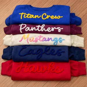 May include: A stack of crewneck sweatshirts in various colors: blue, burgundy, cream, navy, and red. Each sweatshirt features a team name in a contrasting, embroidered script: "Titan Crew," "Panthers," "Mustangs," "Eagles," and "Hawks."