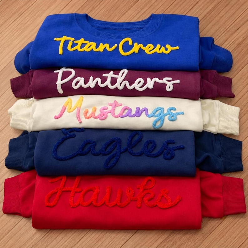 May include: A stack of crewneck sweatshirts in various colors: blue, burgundy, cream, navy, and red. Each sweatshirt features a team name in a contrasting, embroidered script: "Titan Crew," "Panthers," "Mustangs," "Eagles," and "Hawks."