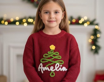 May include: A young person wearing a red knit sweater with a green and yellow Christmas tree design. The name "Amelia" is written in white script below the tree. The sweater has a ribbed texture.