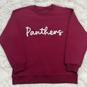 May include: A maroon sweatshirt featuring the word "Panthers" in white, textured lettering. It has a crew neck and ribbed cuffs and hem. The material looks soft and comfortable. The sweatshirt is a classic style.