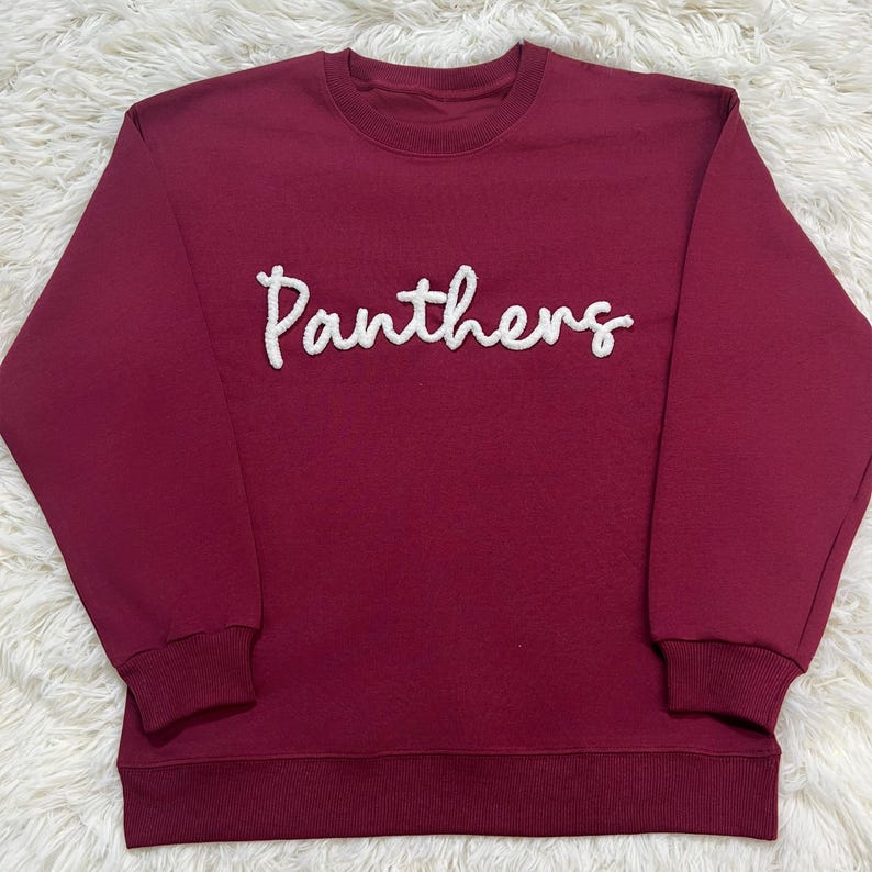 May include: A maroon sweatshirt featuring the word "Panthers" in white, textured lettering. It has a crew neck and ribbed cuffs and hem. The material looks soft and comfortable. The sweatshirt is a classic style.