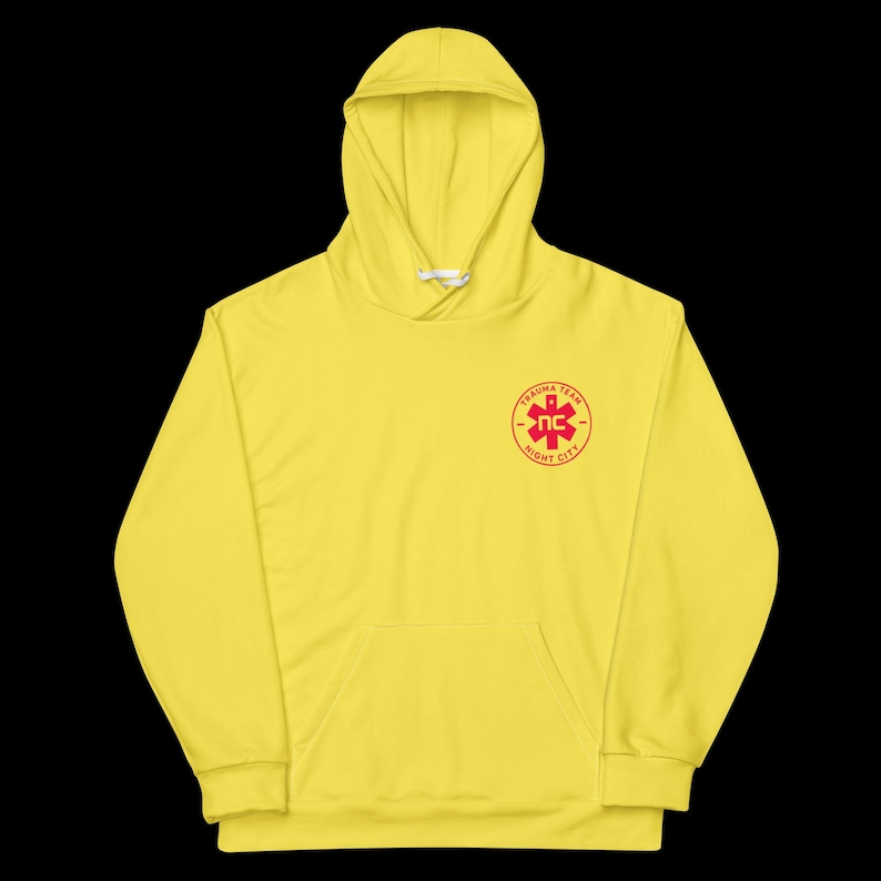May include: A bright yellow hoodie with a front pocket and a drawstring hood. The hoodie features a red emblem on the chest with the text "TRAUMA TEAM" and "NC NIGHT CITY". The garment is made of a soft material.