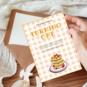 Puede incluir: Una invitación de cumpleaños con un fondo de patrón de cuadros. El texto "TURNING ONE" está en naranja. La invitación incluye la fecha, la hora y el lugar del evento, junto con un contacto RSVP. Ilustración de panqueques con bayas.