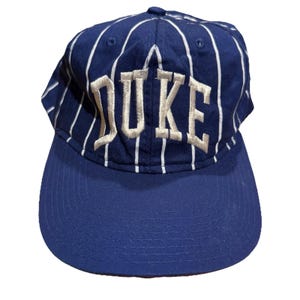 May include: A blue baseball cap with white vertical stripes and the word "DUKE" embroidered in a light tan color. The cap has a curved brim and a six-panel design.