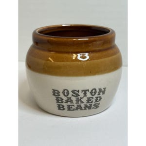 May include: A small, ceramic jar with a brown top and a white base. The words "BOSTON BAKED BEANS" are printed in dark letters on the front of the jar. The jar has a rounded shape and a small opening.