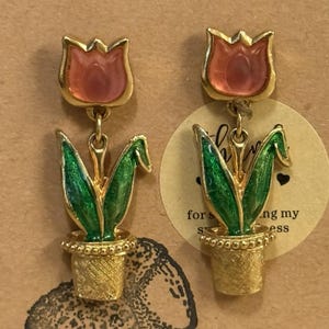 May include: A pair of gold-toned earrings featuring a tulip design. Each earring has a pink tulip flower, green leaves, and a gold pot. The earrings are attached to a post. The earrings are on a brown background.