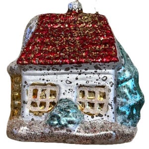 May include: A glass Christmas ornament shaped like a house. The roof is red with glitter, and the house is white with two windows. A small blue tree and silver glitter surround the house.