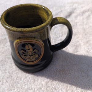 May include: A ceramic mug with a glossy black base and a speckled olive green rim. The front features a circular embossed emblem with a winged helmet and crossed axes, displaying the text Valhalla Java in a raised, tan-colored font.