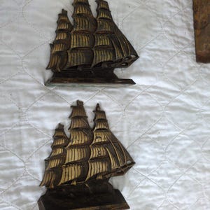 May include: Two antique brass ship sculptures with detailed sails and hulls. The ships have a weathered, aged appearance with gold and brown tones. The sculptures are placed on a white quilted surface, suggesting a nautical or vintage theme.
