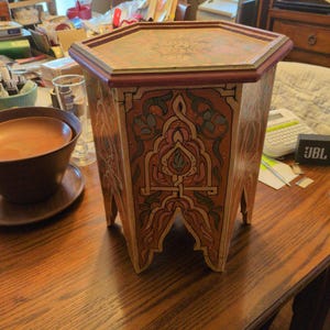 May include: A hexagonal wooden side table featuring a painted finish in shades of orange, red, and green. The table has a flat top with a central floral motif and sides decorated with intricate, symmetrical patterns and arched cutouts at the base.