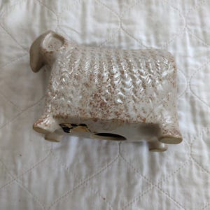 May include: A ceramic sheep figurine in a cream color with brown speckles. The sheep has a textured body, four legs, and a small head with curved horns. The figurine has a coin slot on the bottom, suggesting it is a bank.