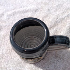 May include: A ceramic mug with a dark, glossy black rim and handle, featuring a light gray interior with visible concentric spiral textures along the inner walls and base. The exterior body displays a circular emblem with a skull graphic.