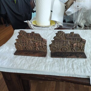 May include: Two bronze-colored bookends featuring a stagecoach design with horses and riders. The bookends are rectangular with detailed relief carvings, and are displayed on a white quilted surface.