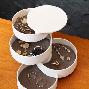 Peut inclure: Un organisateur de bijoux blanc à plusieurs niveaux, avec plusieurs compartiments, chacun doublé de tissu gris. L'organisateur contient diverses boucles d'oreilles, bagues et autres bijoux en or et en argent. Le fond est une surface sombre et texturée.