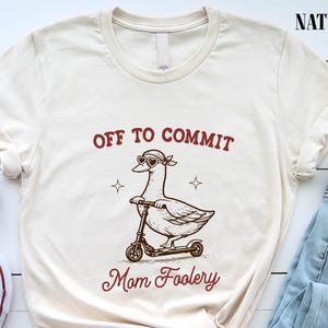 May include: Cream-colored t-shirt with the text "OFF TO COMMIT Mom Foolery" above an illustration of a goose wearing sunglasses and riding a scooter. The shirt has a crew neck and short sleeves. The word "NATURAL" is printed on the top right.