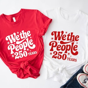 Two t-shirts with the text "We the People 250 Years" printed on them, along with a pair of sneakers and a pair of jeans.