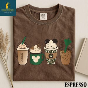 May include: Brown t-shirt featuring four cartoon coffee drinks with whipped cream and Mickey Mouse designs. The shirt has the text "ESPRESSO" at the bottom and a logo in the upper left corner.