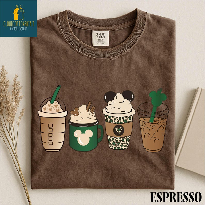 May include: Brown t-shirt featuring four cartoon coffee drinks with whipped cream and Mickey Mouse designs. The shirt has the text "ESPRESSO" at the bottom and a logo in the upper left corner.