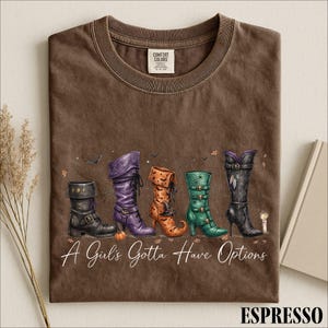 May include: A brown t-shirt featuring a graphic of five distinct boots in black, purple, orange, green, and black. The text "A Girl's Gotta Have Options" is printed in white script below the boots. The shirt has a Comfort Colors brand label.
