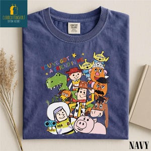 K&ouml;nnte beinhalten: Marineblaues T-Shirt mit einem farbenfrohen Aufdruck von Toy Story-Charakteren und dem Schriftzug "YOU'VE GOT A FRIEND IN ME". Das gefaltete Shirt hat ein Comfort Colors-Etikett. Das Wort "NAVY" ist unten aufgedruckt.