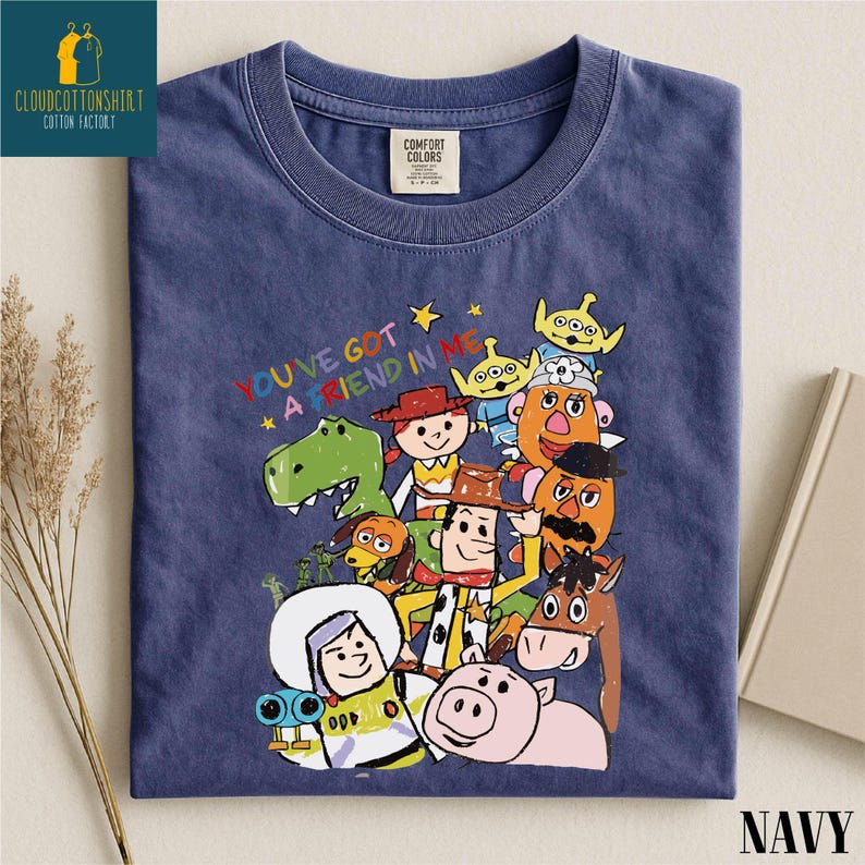 K&ouml;nnte beinhalten: Marineblaues T-Shirt mit einem farbenfrohen Aufdruck von Toy Story-Charakteren und dem Schriftzug "YOU'VE GOT A FRIEND IN ME". Das gefaltete Shirt hat ein Comfort Colors-Etikett. Das Wort "NAVY" ist unten aufgedruckt.