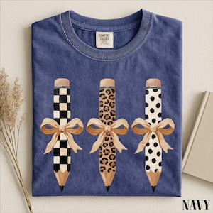 A navy blue t-shirt with a graphic design of three pencils with bows, each with a different pattern: a checkered pattern, a leopard print pattern, and a polka dot pattern.