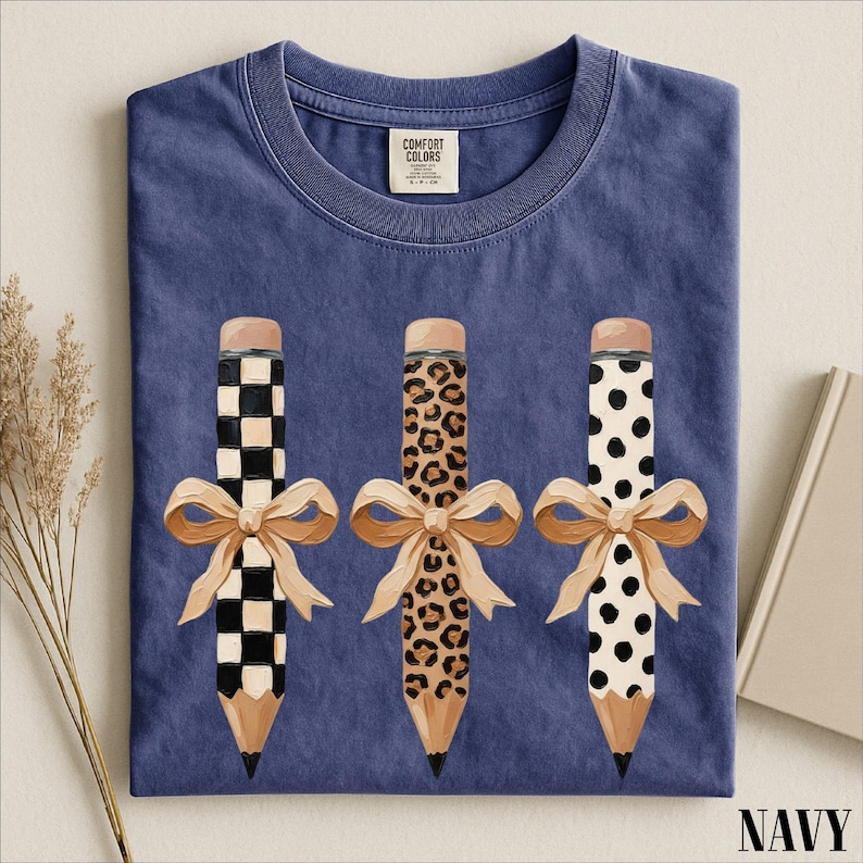 A navy blue t-shirt with a graphic design of three pencils with bows, each with a different pattern: a checkered pattern, a leopard print pattern, and a polka dot pattern.
