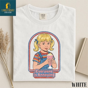 A white t-shirt with a graphic design depicting a young girl with blonde hair and a straw, holding a milkshake, with the text "Everyone is Annoying" below the image.