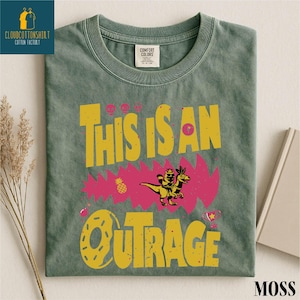 a green t-shirt with a colorful graphic design, including a dinosaur, flowers, and the text "This is an OTRAGE".