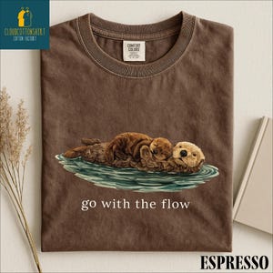 a brown t-shirt with a printed design of two otters swimming together in a body of water, accompanied by the text "go with the flow".