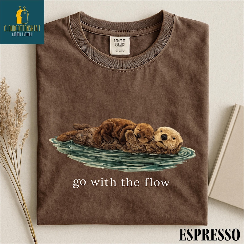 a brown t-shirt with a printed design of two otters swimming together in a body of water, accompanied by the text "go with the flow".