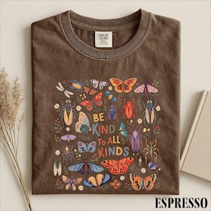 a brown t-shirt with a colorful graphic design of various insects and butterflies, along with the text "Be Kind to All Kinds" printed on it.