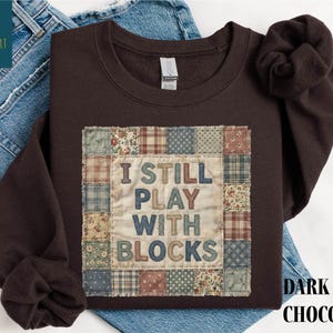May include: Dark chocolate-colored sweatshirt with a patchwork design featuring the text "I STILL PLAY WITH BLOCKS." The patchwork includes various floral and plaid patterns. The sweatshirt is displayed on a pair of blue jeans.