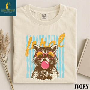a white t-shirt with a graphic design of a raccoon wearing sunglasses and blowing a bubble gum bubble.