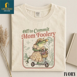May include: Ivory-colored t-shirt with the phrase "Off to Commit Mom Foolery" in green and red. The shirt features a whimsical illustration of a frog riding a scooter, wearing a dress and hat, with a basket of flowers.