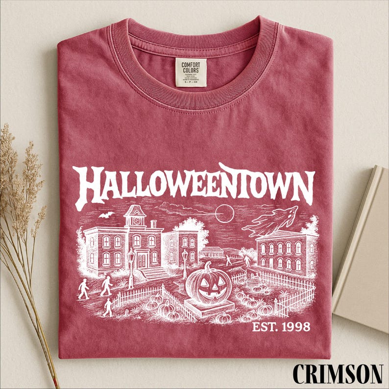 A red t-shirt with a Halloween-themed graphic design, including a pumpkin patch, buildings, and a clock tower, along with the text "Halloweentown" and "EST. 1998".