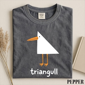 May include: A charcoal gray crew neck t-shirt featuring a white triangular bird graphic with an orange beak and legs. Below the bird, the word "triangull" is printed in white. The shirt has a Comfort Colors label at the inner neckline.