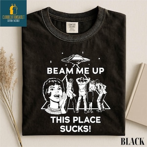 May include: Black t-shirt with a retro graphic design featuring a UFO, stars, and the phrase "BEAM ME UP THIS PLACE SUCKS!". The design includes a woman and several people with their arms raised. The shirt has a crew neck.