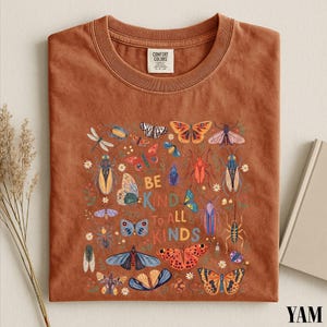 a brown t-shirt with a colorful graphic design of various insects and butterflies, along with the text "Be kind to all kinds".