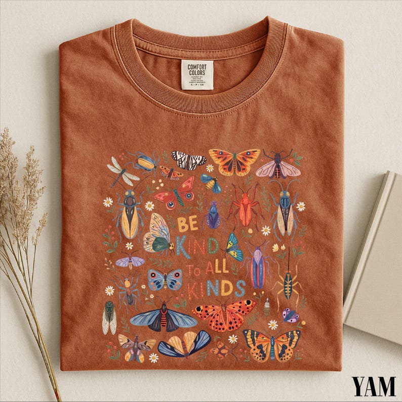 a brown t-shirt with a colorful graphic design of various insects and butterflies, along with the text "Be kind to all kinds".
