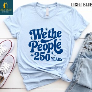 A light blue t-shirt with a graphic design that says "We the People 250 Years" printed on it, along with some jeans and white sneakers.