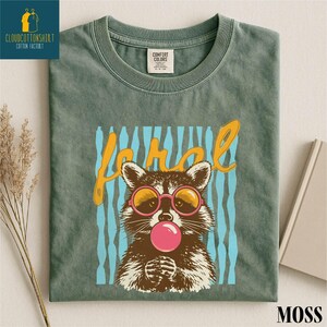 a green t-shirt with a graphic design of a raccoon wearing sunglasses and blowing a bubble gum bubble.